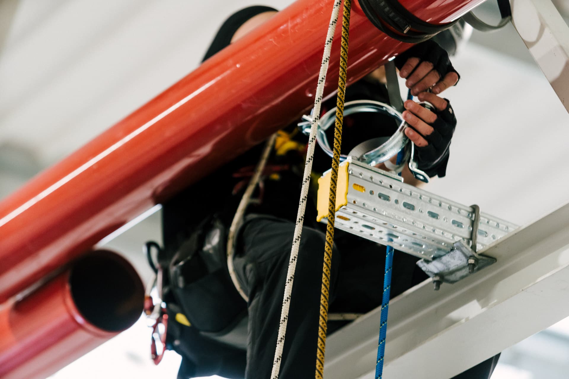 Worker in safety gear climbing a ladder, inspecting large red pipes, with ropes and equipment visible.