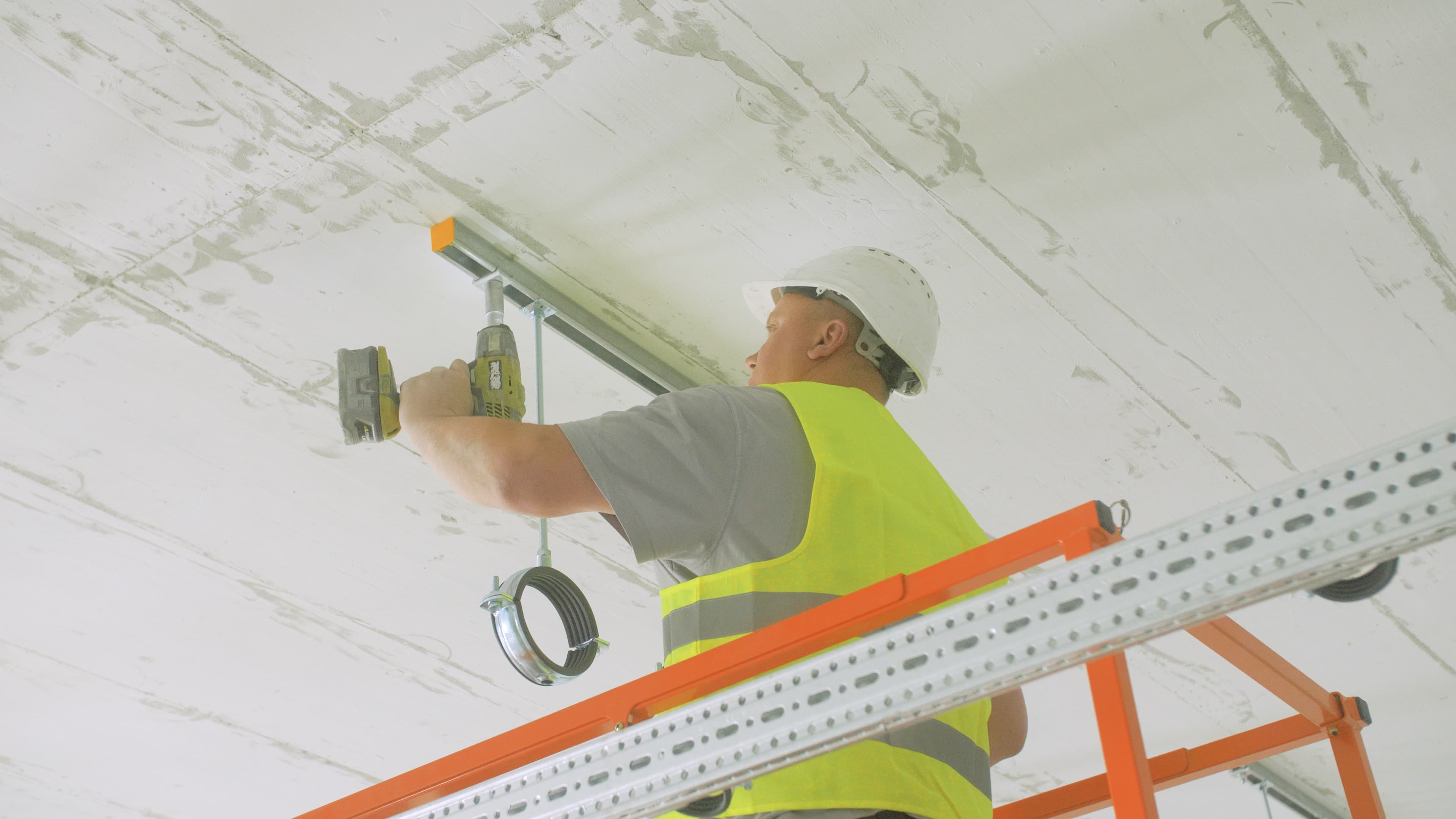 Worker in a hard hat and safety vest uses a power drill to install a metal siConnect on a ceiling, standing on an orange scaffold.