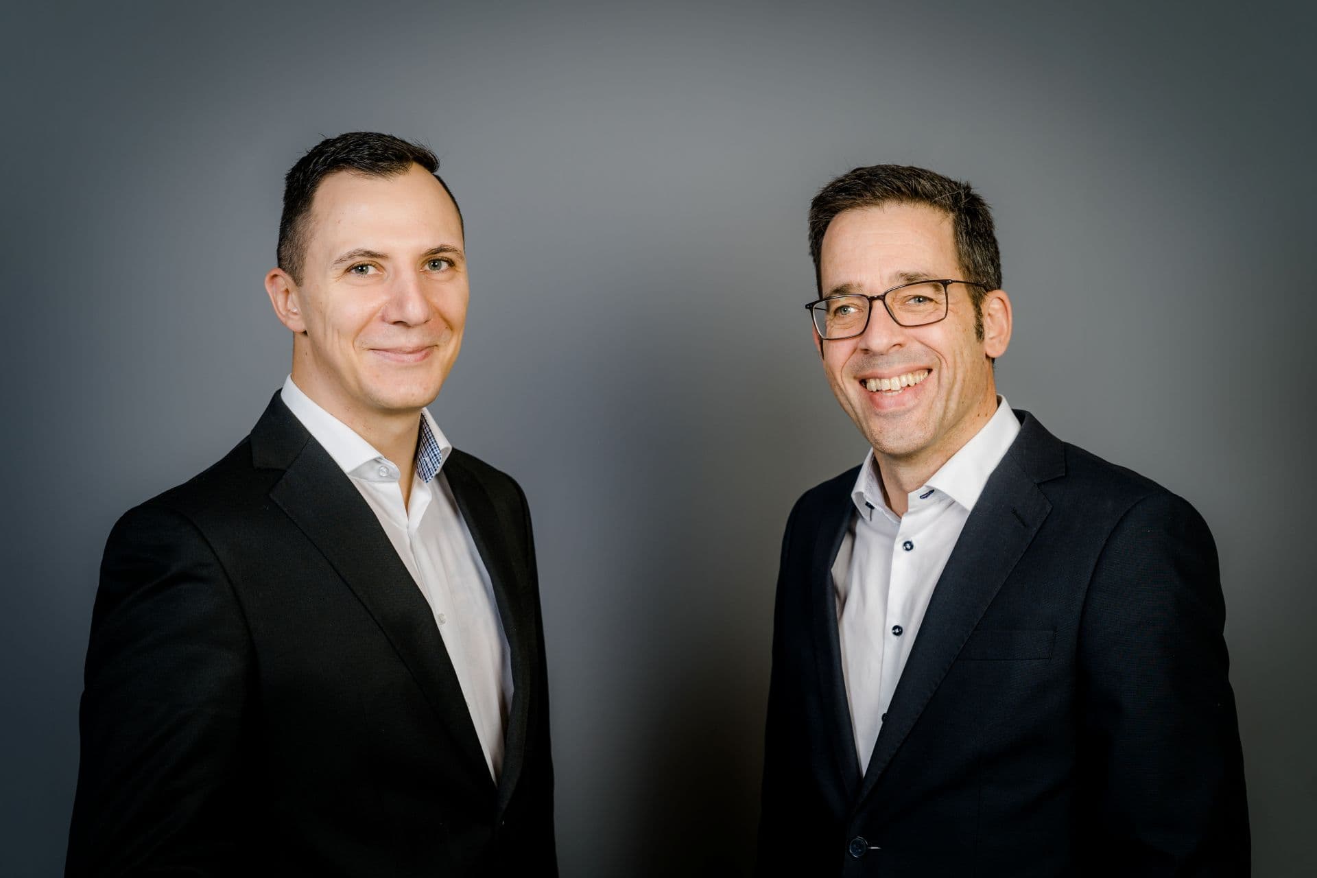 Two men in business attire stand against a gray background, smiling at the camera.